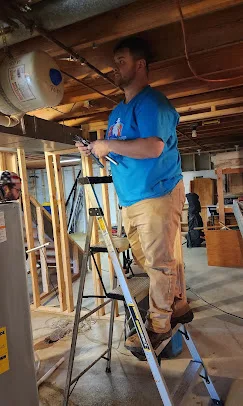 Patriot AC tech in a uniform T-shirt standing on a ladder with a tool in hand looking at an indoor air conditioning system