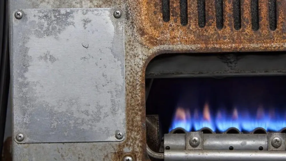 Close-up of a corroded, rust-covered furnace panel with blue burner flames visible through the open burner compartment