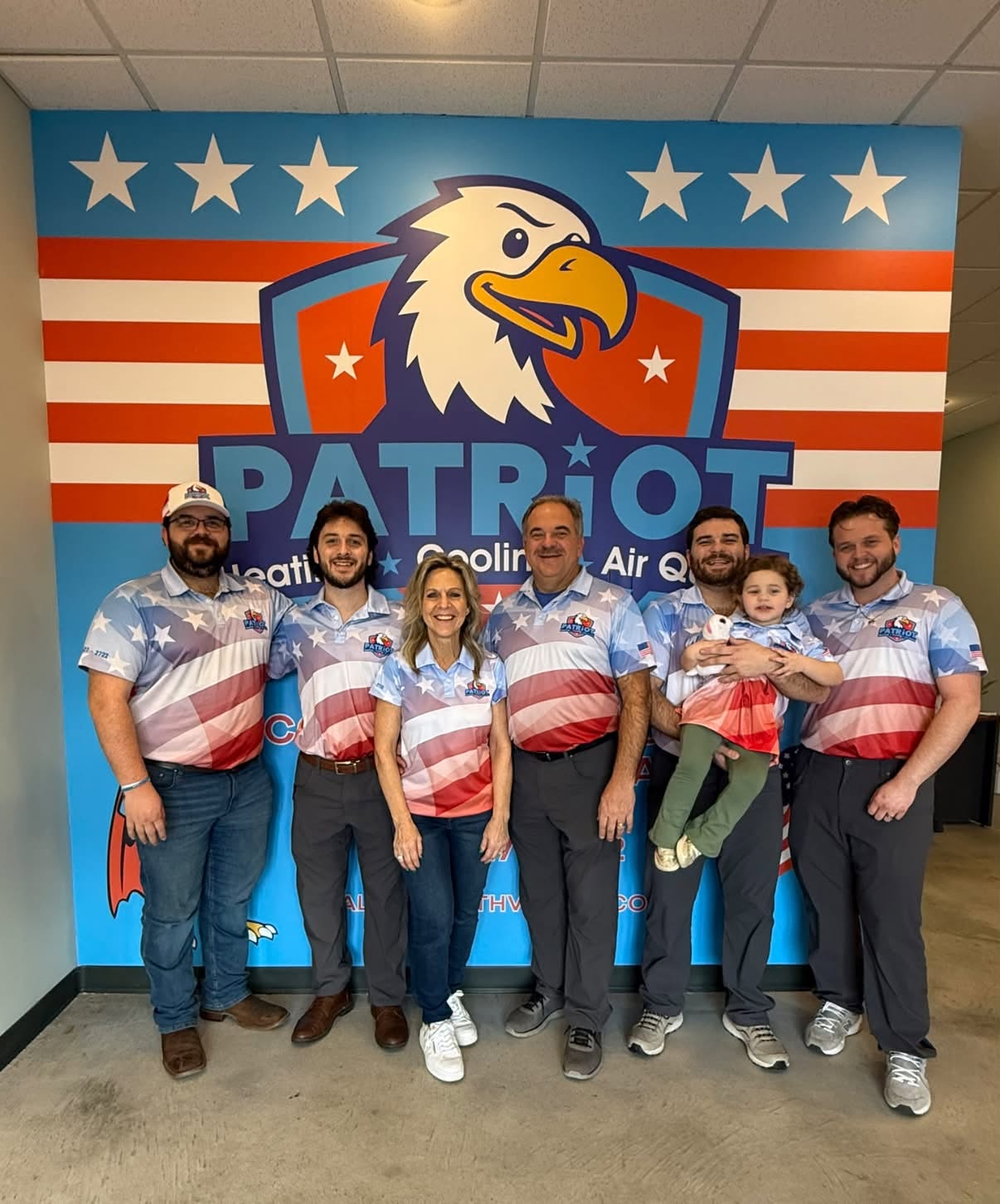 The Patriot Heating, Cooling & Air Quality family team in matching flag polos in front of the Patriot eagle wall at their office